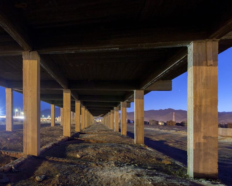 View along the underside of an unfinished concrete building at dusk, pillars receding toward town lights and desert mountains, Egypt