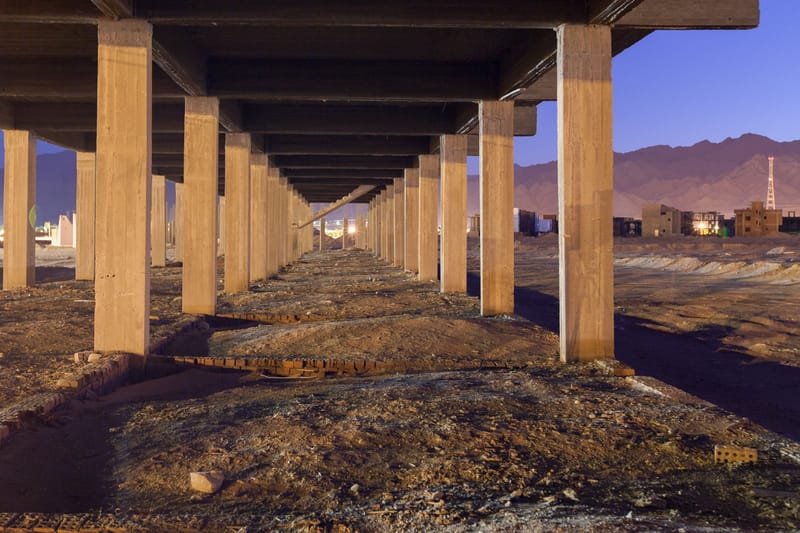 Concrete pillars of an abandoned, unfinished building at dusk, town lights and mountains on the horizon