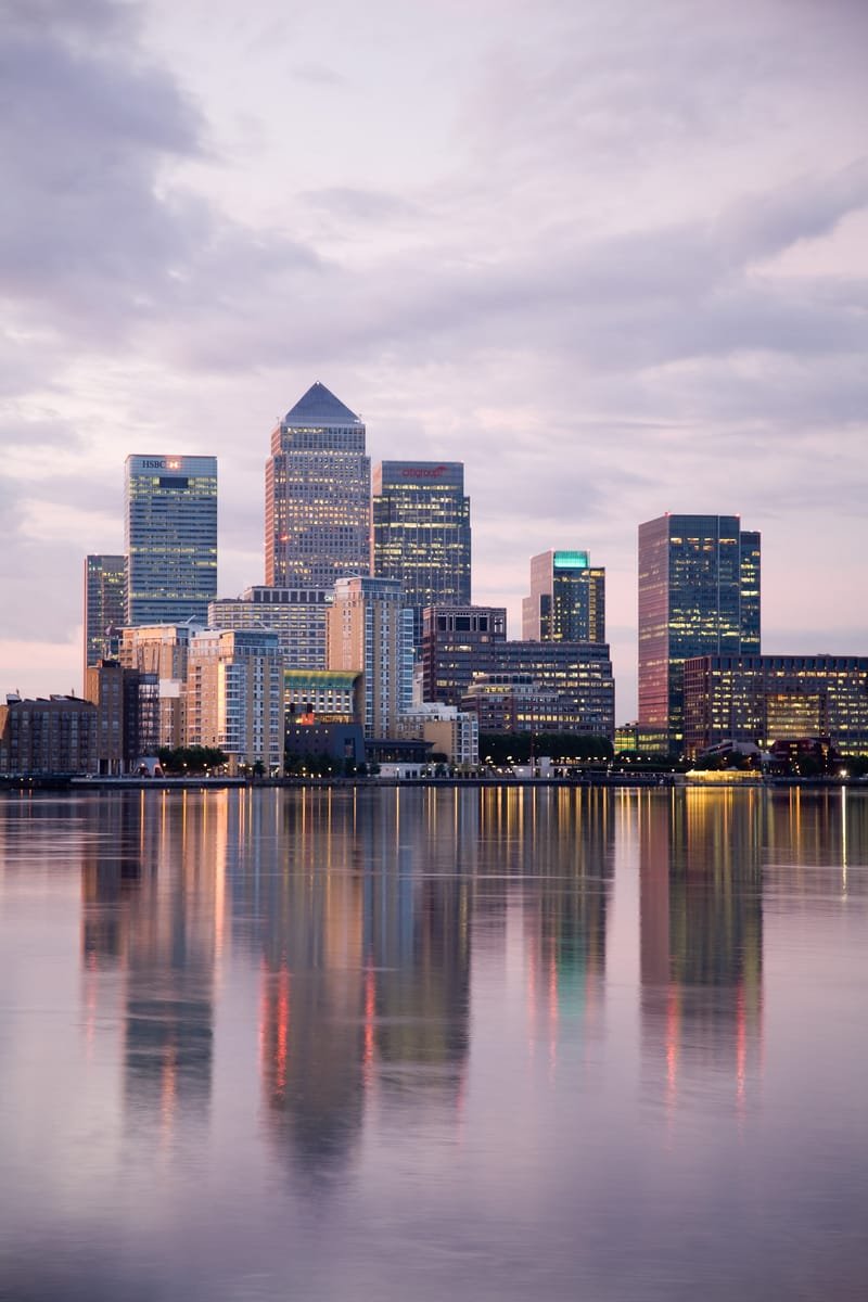 The Canary Wharf cluster reflected in calm water at sunrise under a pastel sky