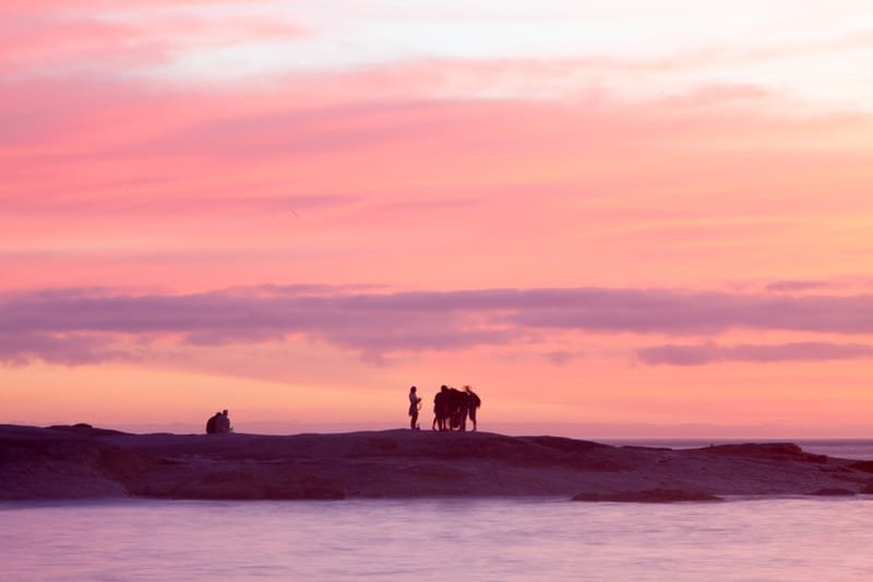 A small group silhouetted on a sea rock under layered pink clouds at dusk, Rio de Janeiro